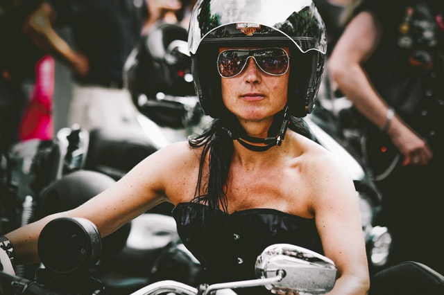 Žena v čiernom korzete, so slnečnými okuliarmi a  čiernou prilbou na hlave sedí na motorke